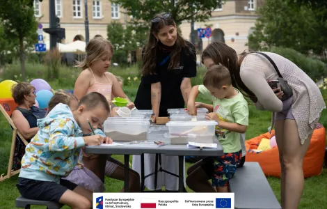 Grupa osób (dwie osoby dorosłe i czwórka dzieci) przy stole piknikowym podczas warsztatów archeologicznych. Jedna z dorosłych osób pochyla się nad jednym z dzieci przypatrując się jego twórczości. W tle plac Wolności oraz starsza kobieta w okularach przeciwsłonecznych siedząca na leżaku. 