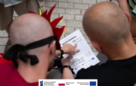 Zbliżenie na kartkę z napisem „Szyfr Feniksa” oraz wskazujący na nią palec. 