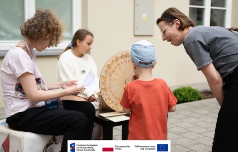 Dwie kobiety oraz dwójka dzieci podczas quizu etnograficznego, na pierwszym planie koło z literami i cyframi wykonane z drewna. 