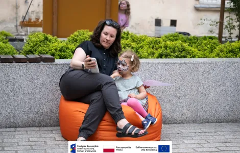 Ubrana na ciemno kobieta z okularami przeciwsłonecznymi założonymi jak opaska na włosy wraz z małą dziewczynką, która ma pomalowaną twarz we wzór kota siedzą na pomarańczowej ogromnej pufie i robią sobie zdjęcie selfie