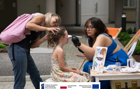 Kobieta wraz z dziewczynką uczesaną w kucyk podczas malowania twarzy. Po prawej malująca z ciemnymi włosami ubrana w kobaltową sukienkę. 