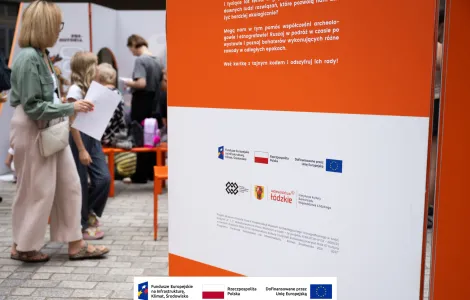 Na pierwszym planie jedna ze ścianek wystawy informująca o przyznanym dofinansowaniu z Unii Europejskiej oraz logotypami. W tle przechadzający się zwiedzający. 