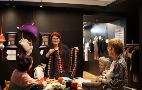 trzy kobiety podczas warsztatów tekstylnych, dwie siedzą po przeciwnych stronach stołu, jedna z nich stoi na środku i coś im tłumaczy