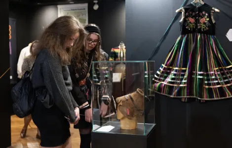 Dwie młode kobiety oglądają w muzeum eksponat - czapkę prezentowaną w szklanej gablocie. W tle na ścianie wisi kolorowa, haftowana sukienka ludowa.