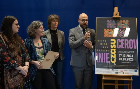 trzy młode kobiety oraz mężczyzna w garniturze i okularach stojący obok sztalugi, na której wyeksponowano plakat wystawy