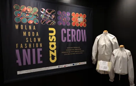 zdjęcie przedstawiające banner z tytułem wystawy, utrzymany w ciemnej kolorystyce z żółtymi oraz fioletowymi napisami oraz logotypami unijnymi. Po prawej dwa manekiny ubrane w stroje ludowe