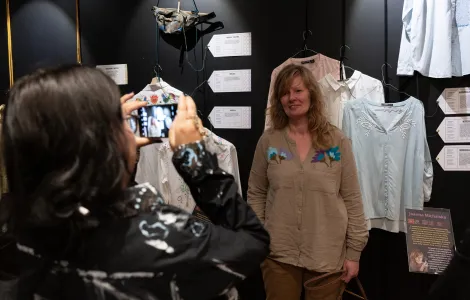 młoda kobieta robiąca zdjęcie telefonem drugiej młodej kobiecie na tle tekstylnych eksponatów zawieszonych na ścianie 