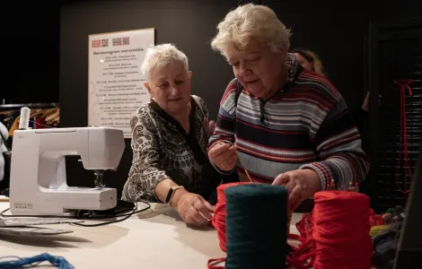 dwie starsze kobiety przy stole z maszyną do szycia wybierające kawałki tkanin
