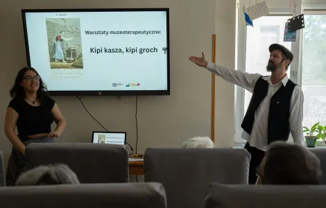 Na zdjęciu po lewej stronie stoi młoda śmiejąca się kobieta. Po prawej stronie stoi mężczyzna z brodą, w białej koszuli, czarnej kamizelce i czapce z daszkiem. Wskazuje ręką na ekran z napisem Kipi kasza, kipi groch.