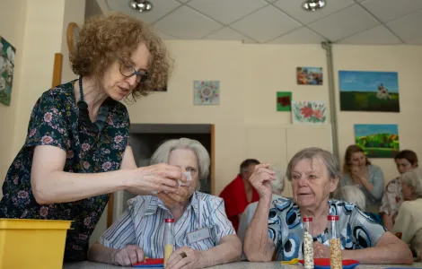 Przy stole siedzą dwie starsze panie. Przed nimi stoją fiolki z różnymi ziarnami. Po lewej stronie stoi starsza kobieta.