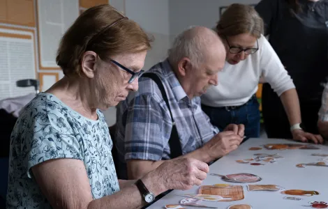 Przy stoliku siedzą starsi kobieta i mężczyzna. Przed nimi na stole leżą różne obrazki, które oni oglądają. Obok mężczyzny stoi kobieta  w okularach.