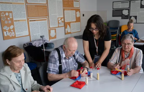 Przy stole siędzą starsze dwie kobiety i starszy mężczyzna. Między nimi stoi młoda kobieta. Starsze osoby dotykają leżących na stole kolorowych woreczków.