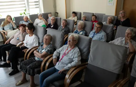 Na zdjęciu widać sporą grupę osób, głównie starszych. Siedzą w dużych, szarych fotelach. Wszyscy patrzą w jednym kierunku.