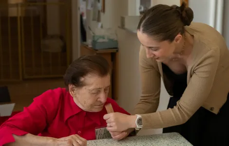 W centrum zdjęcia widać starsza kobietę w czerwonej bluzce. Wącha ona fiolkę. Fiolkę trzyma młoda kobieta w beżowej bluzce, z czarnymi włosami spiętymi w koczek.
