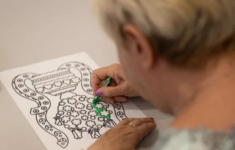 Starsza kobieta koloruje na zielono obrazek. Najbardziej widoczny jest czarno - biały obrazek przedstawiający koronę ślubną.