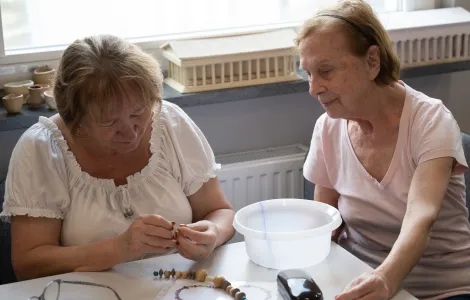 Dwie starsze panie siedzą przy stole. Przed nimi na stole stoi miseczka i leżą korale. Kobieta po lewej nawleka koralik na sznurek.