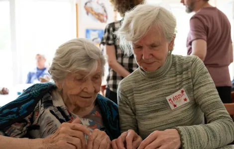 Dwie starsze, siwe kobiety siedzą obok siebie przy stole. Są wpatrzone w korale leżące przed nimi.