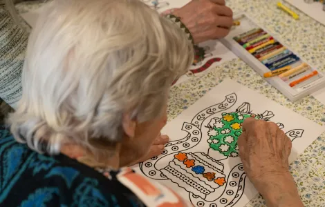 Starsza kobieta w niebieskim swetrze koloruje obrazek korony ślubnej, używając pasteli olejnych.