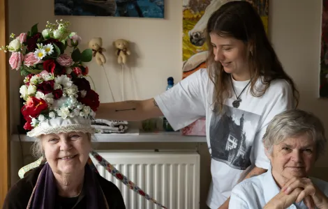 Z lewej strony siedzi starsza, uśmiechnięta kobieta. Ma na głowie bardzo kolorową, pełną kwiatów koronę ślubną. Obok niej stoi młoda kobieta w białej bluzce i siedzi starsza kobieta, która lekko się uśmiecha.