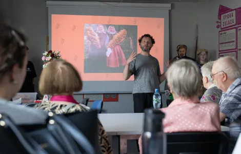 Grupa starszych osób siedzi przy stole i patrzy na ekran. Na ekranie widac kobiety w strojach ludowych z ogromnej wielkości pieczywem. Przed ekranem stoi mężczyzna i gestykuluje.