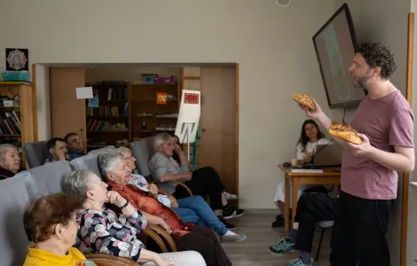 Po prawej stronie stoi mężczyzna w średni  wieku. W rękach trzyma dwa chleby. Przed nim na fotelach siedzi grupa starszych osób.