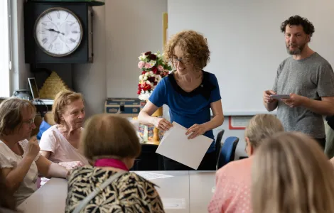 Grupa starszych kobiet siedzi przy stole. Obok stołu stoi kobieta i mężczyzna. Kobieta trzyma w ręku białe kartki.