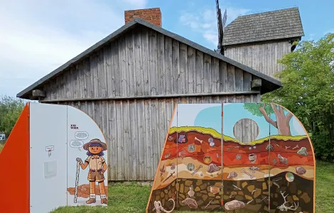 zdjęcie przedstawia ścianki wystawy mobilnej na tle drewnianej zabudowy stodoły. Na jednej ze ścianek widzimy postać archeolożki, na drugiej przekrój warstw ziemi, w której poukrywane są zabytki archeologiczne