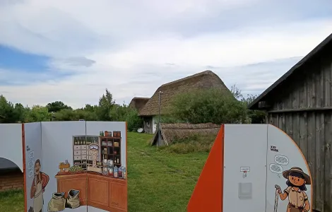 na pierwszym planie dwie ścianki wystawy mobilnej, na jednej z nich wnętrze sklepiku oraz wąsaty mężczyzna, na drugiej archeolożka. W tle zabudowania skansenu