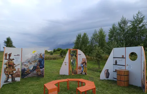 na środku zdjęcia widzimy pomarańczową ławeczkę w kształcie ośmioboku, która ustawiona jest pomiędzy ściankami wystawy mobilnej. W tle zielone drzewa oraz pochmurne niebo