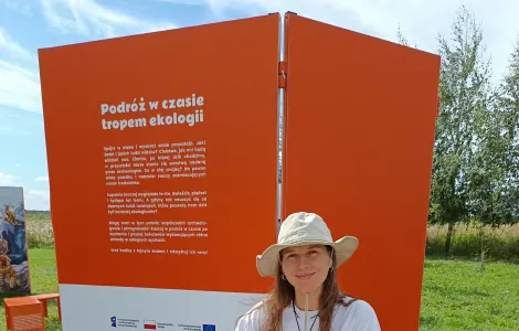 Młoda kobieta w kapeluszu siedząca przy drewnianym stole, na którym stoją pojemniki z piaskiem i szkła powiększające. W tle ścianka wystawowa z napisem "Podróż w czasie tropem ekologii"