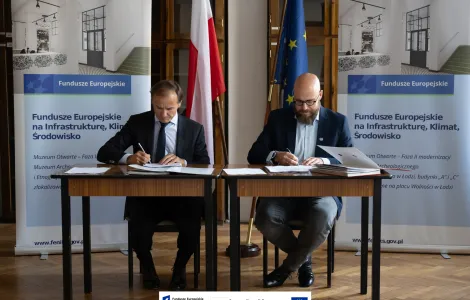 dwóch mężczyzn siedzących przy niewielkich stołach podpisujących dokumenty. W tle flaga Polski i Unii Europejskiej