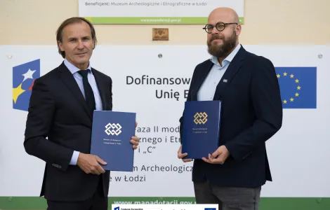 dwóch mężczyzn na tle tablicy informacyjnej z logotypami unijnymi, obaj trzymają granatowe teczki 