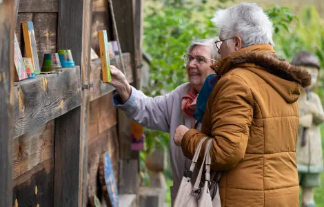 Dwie starsze kobiety ubrane w kurtki patrzą się na ścianę drewnianego domu, na której ułożono kolorowe doniczki i obrazki. Jedna z kobiet trzyma w ręku taki obrazek.