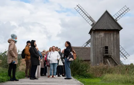 Grupa osób ubranych w kurtki stoi na drodze zbudowanej z drewnianych desek. Z prawej  strony stoi młoda kobieta, która przy ustach ma mikrofon. Za kobietą widać duży, drewniany wiatrak.