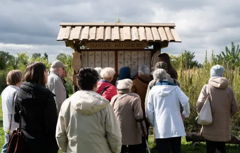 Pod tablicą z napisem Skansen naturalnie stoi duża grupa ludzi. Widzimy ich tyłem. Są ubrani w kurtki, wokół nich pozostaje odkryta polna przestrzeń.