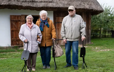 Dwie starsze kobiety i starszy mężczyzna stoją wspierając się o czarne laski z siedziskami. Za nimi widać białą chałupę z dachem ze słomy.