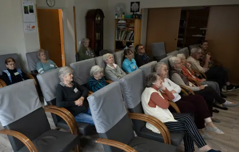 W sali na fotelach siedzi grupa starszych osób. Wszyscy patrzą przed siebie w jednym kierunku.