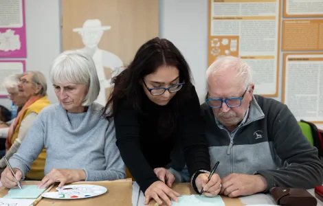 Starsza kobieta i starszy mężczyzna siedzą przy stole. Między nimi stoi młoda kobieta. Rysuje ona coś na kartce. Na stole leżą palety z farbami.
