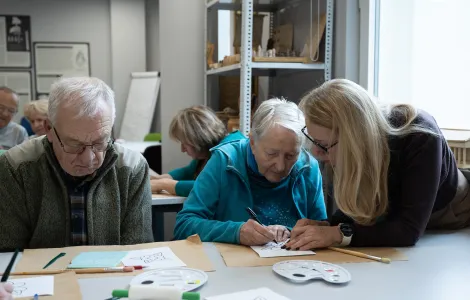 Starsi kobieta i mężczyzna siedzą przy stole. Przed nimi kartki i palety z farbami. Przy starszej kobiecie stoi kobieta z długimi włosami. 